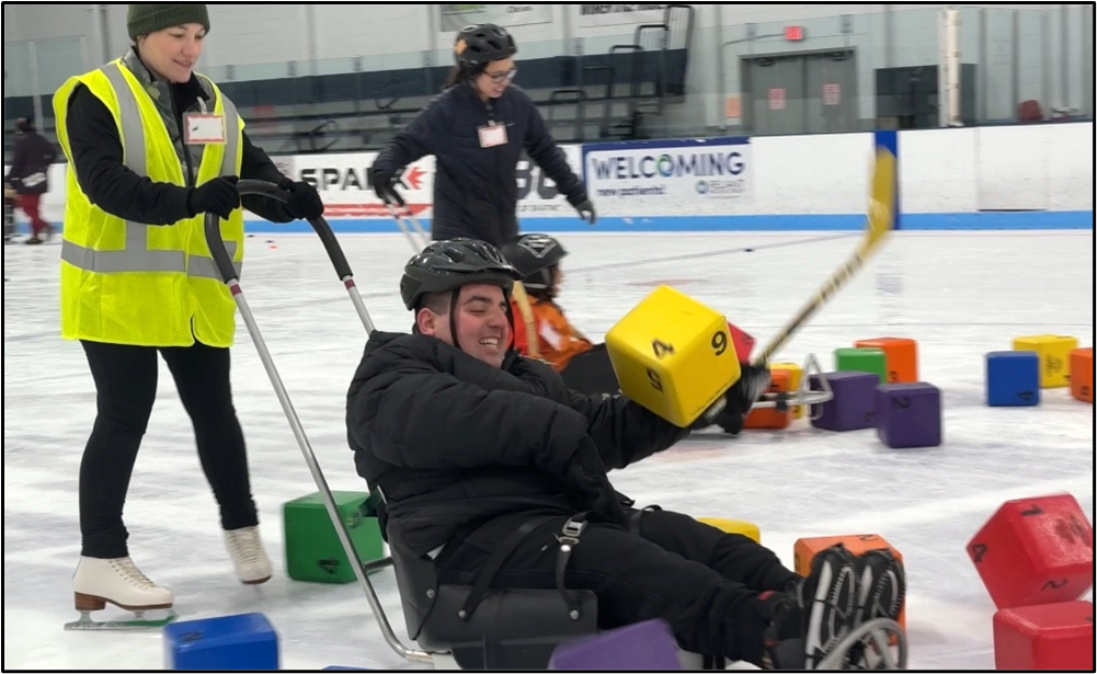 A person using an ice sled laughs while being pushed through foam blocks on the ice. He is trying to hit a block with a hockey stick.
