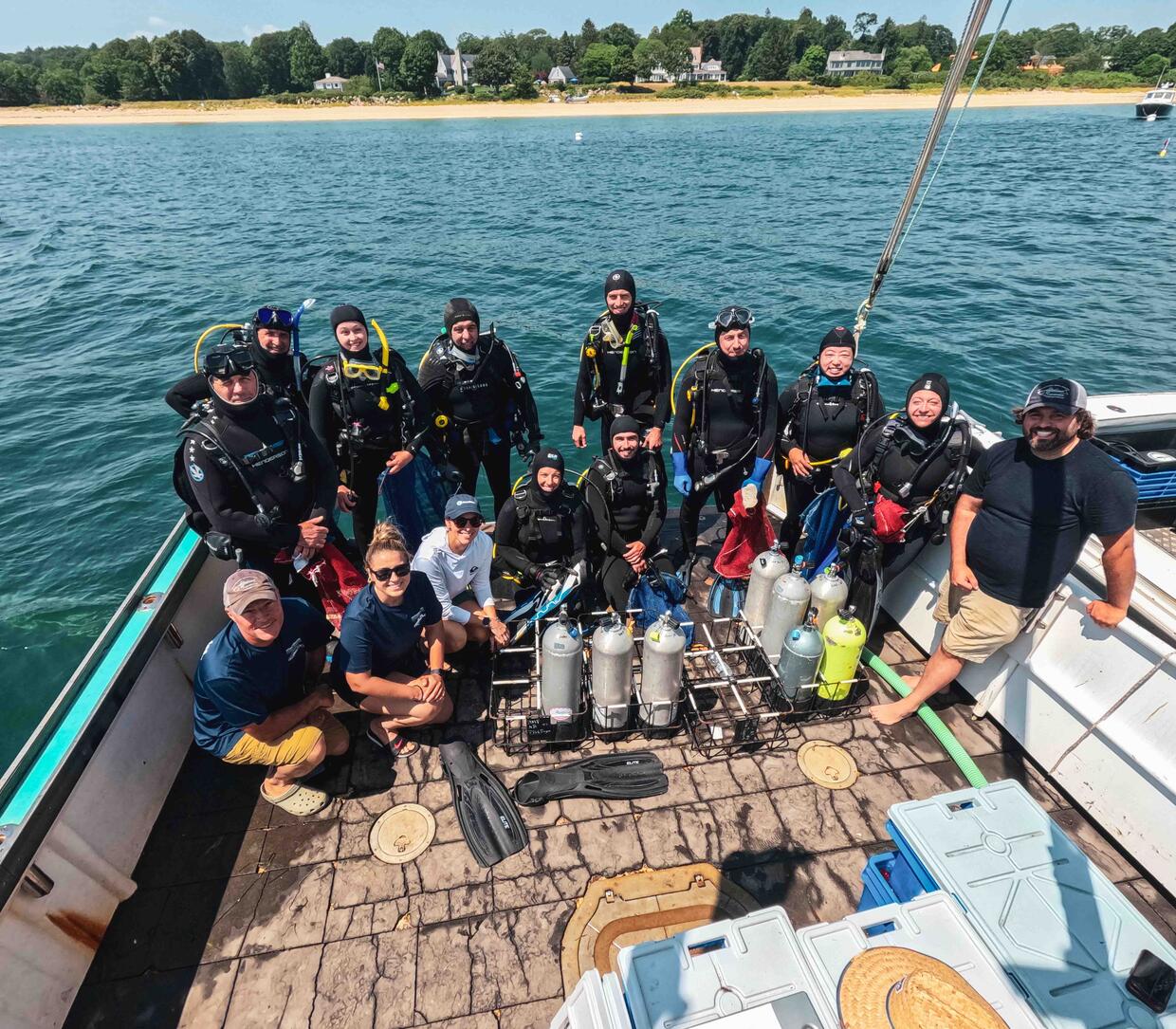A group of 14 scientific divers and support staff stand at the back of a vessel, some wearing dive suits, preparing for their next dive. 
