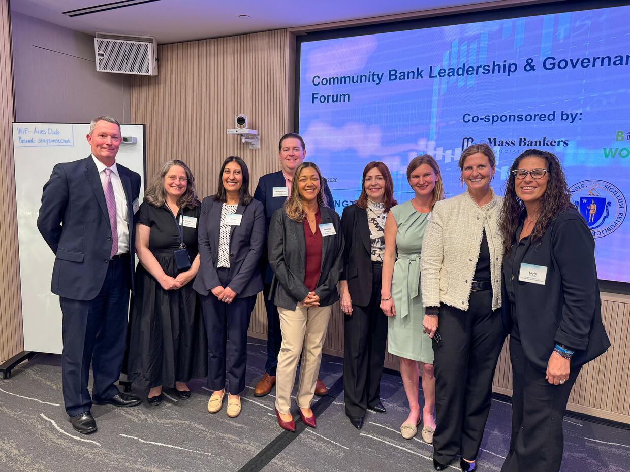 A group of nine people stand in a conference room in front of a screen reading "Community Bank Leadership & Governance Forum.” 