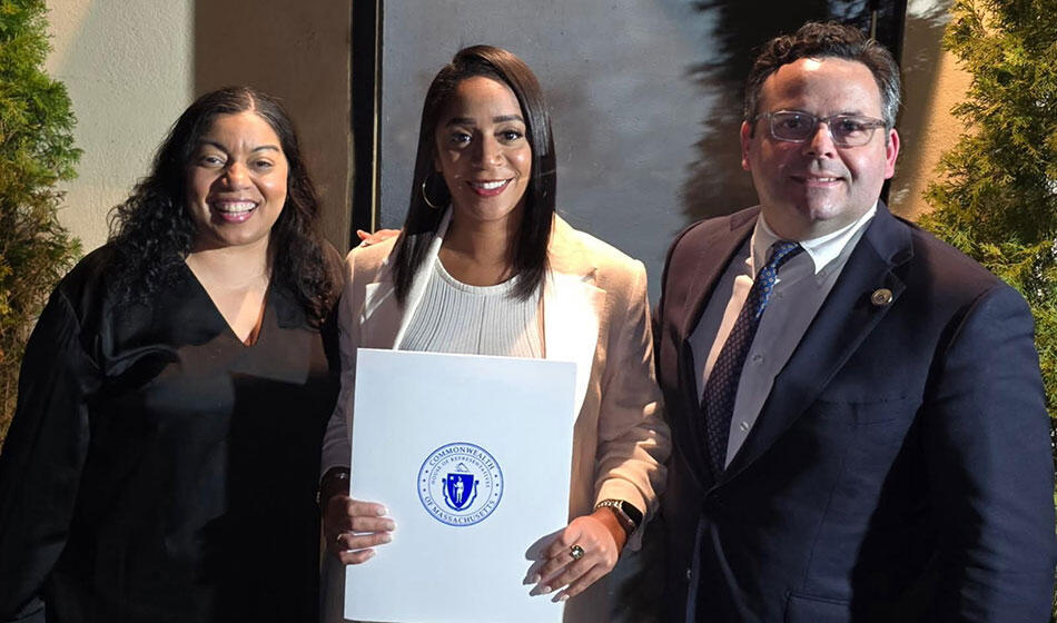 Three people standing outdoors with the person in the middle holding an award.