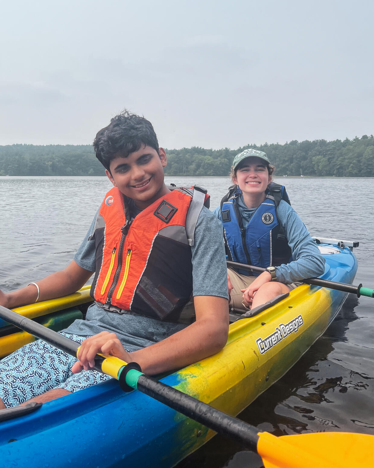 Two people in a tandem kayak on a lake, smile for a photo while holding paddles. 