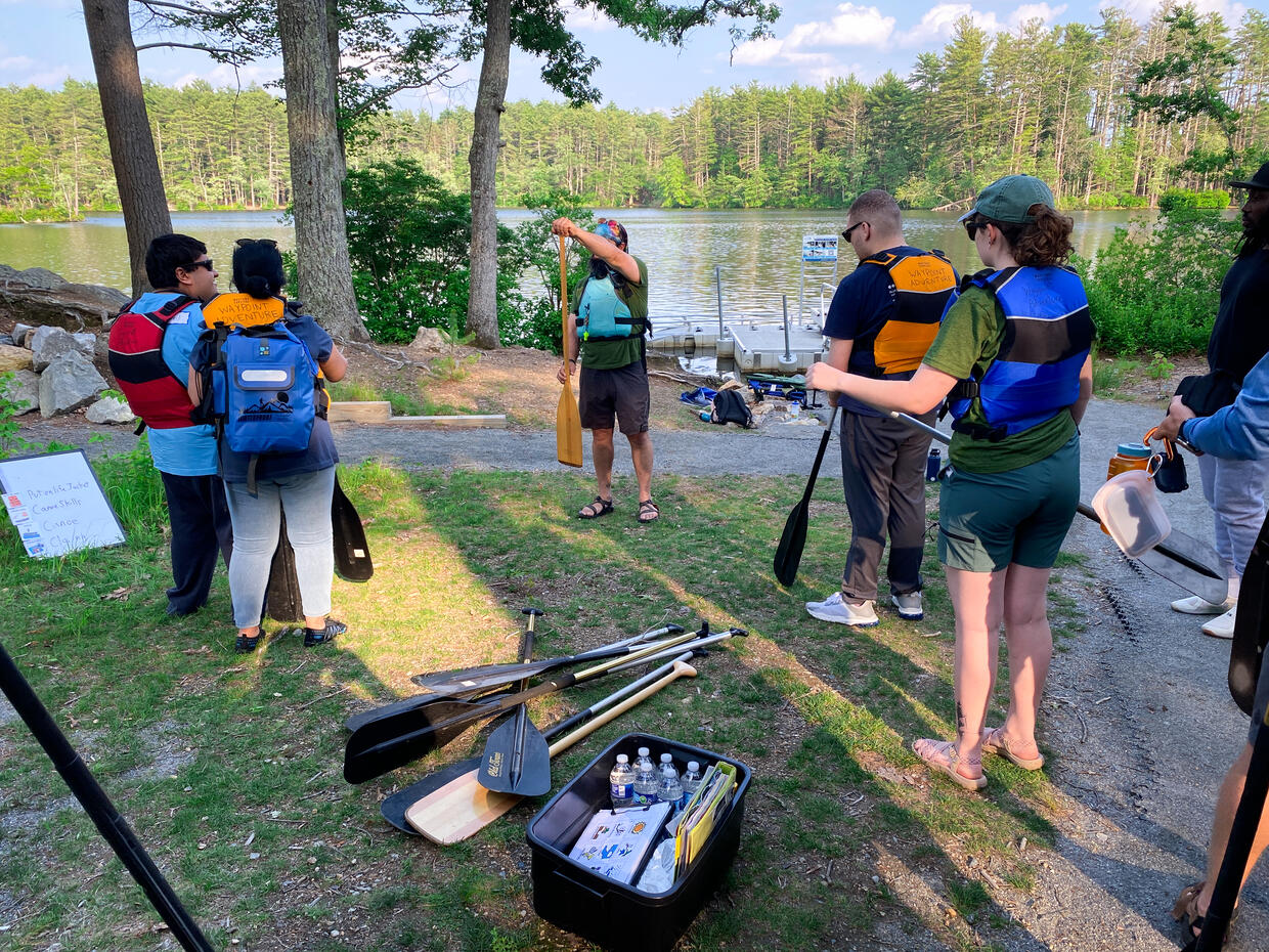 Program staff show a group of people on shore  how to hold an oar. 