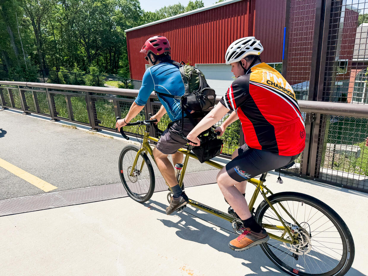 Two people on a tandem bike peddle along a paved bike trail. 