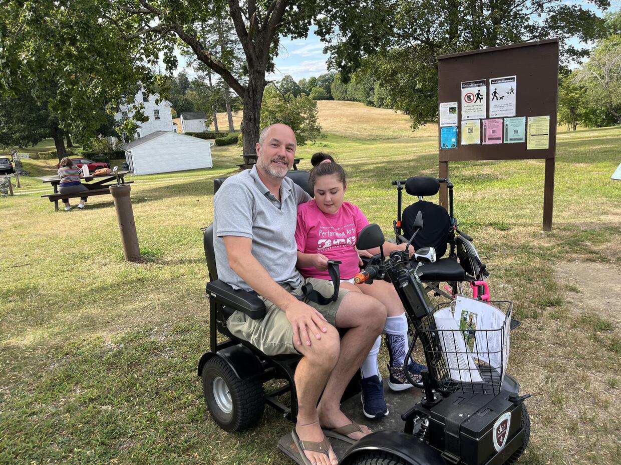 Father and daughter smile for the camera as sit on a dual seat mobility scooter in a park. 