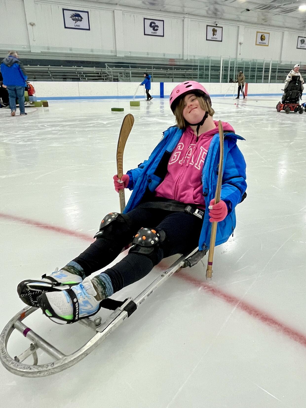 Person seated on ice sled holding adaptive hockey sticks smiles for the camera. 