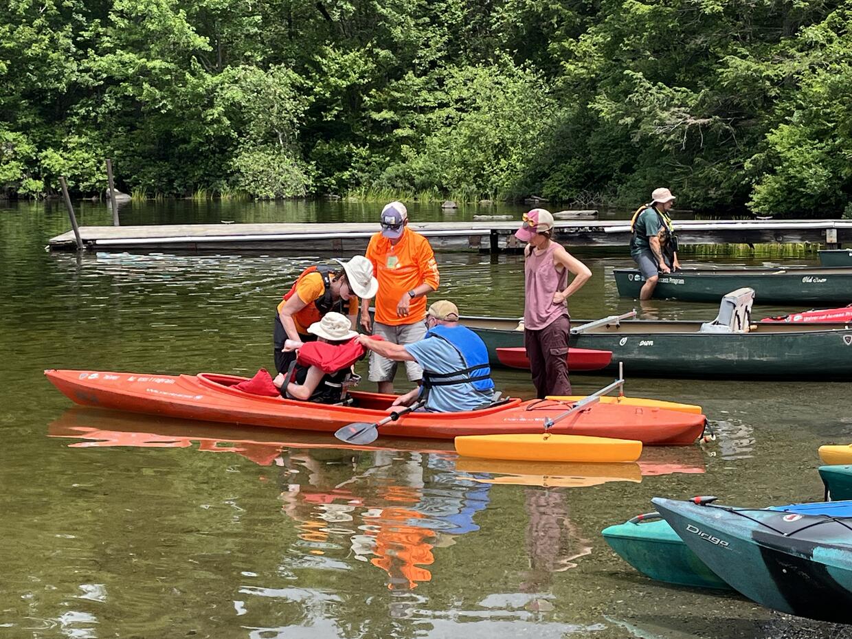 Program staff make adjustments for a person seated in an adaptive seat in a tandem kayak equipped with outriggers. 