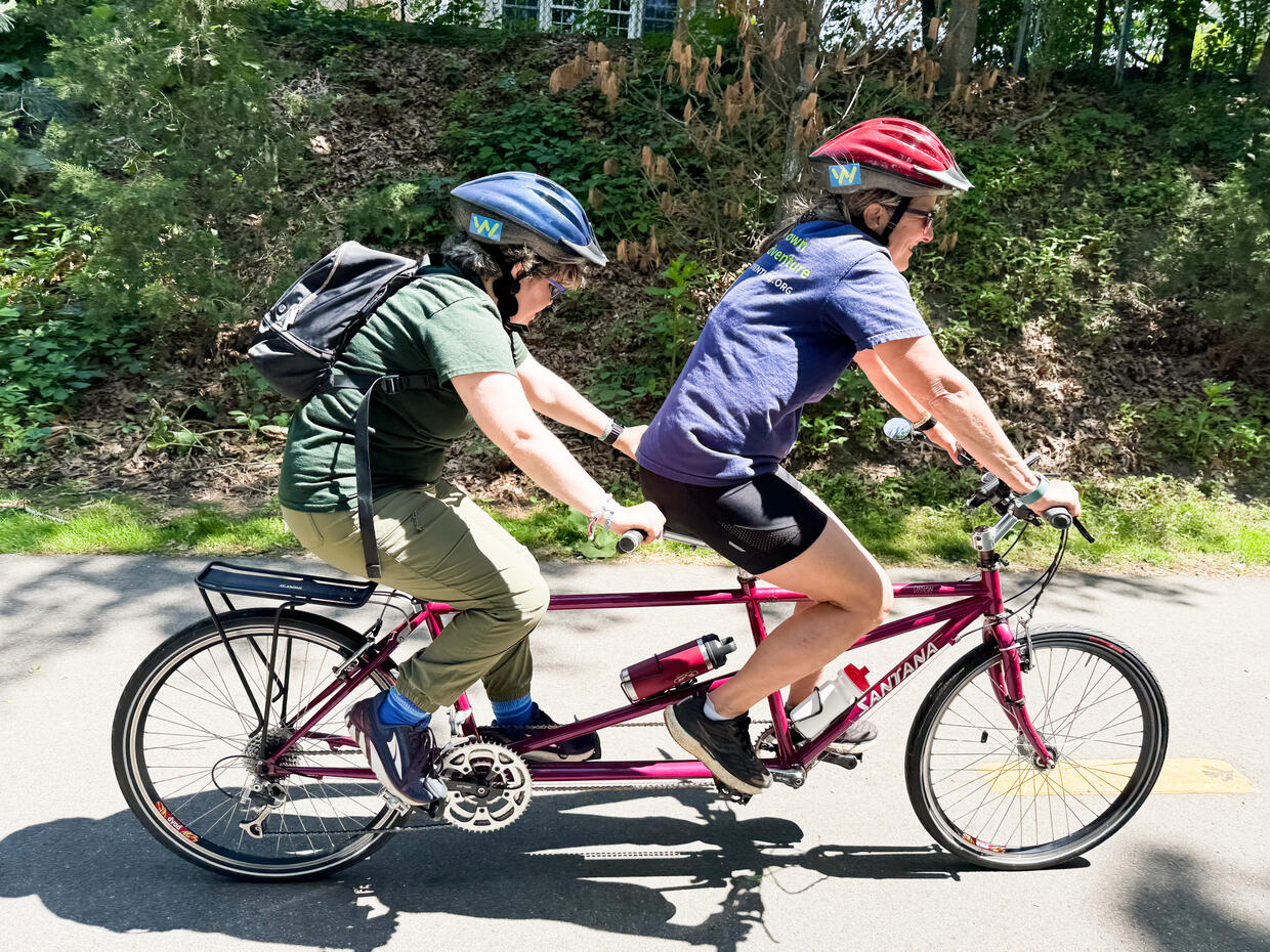 Two people riding a tandem bike peddle along a paved bike trail. 