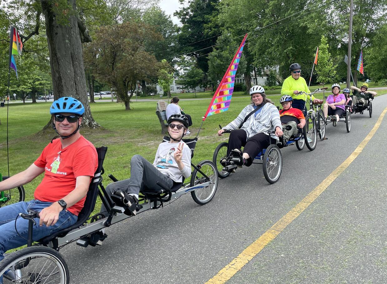 A line of people riding a variety of adaptive cycles on a paved bike trail stop and smile for a photo.