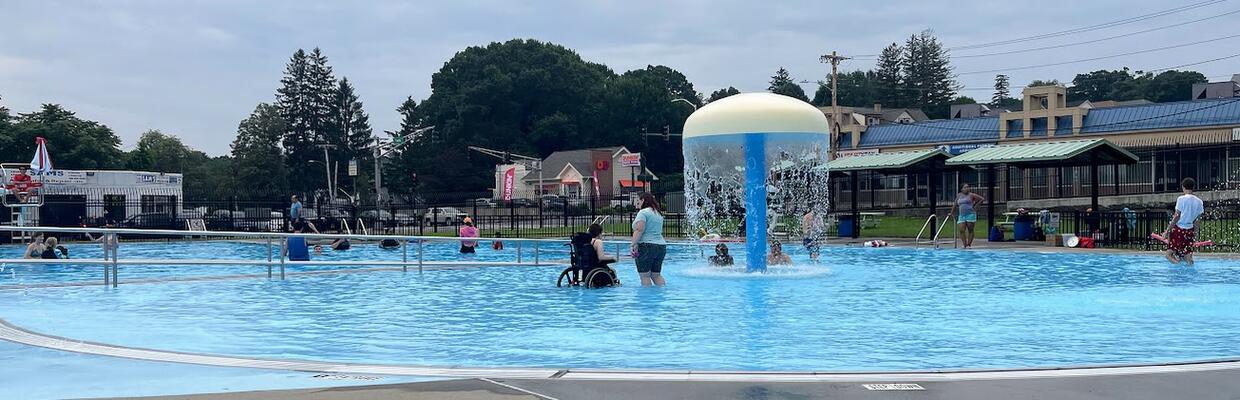 A group of people are playing in a pool equipped with an accessible ramp and splash pad. One person is using their wheelchair in the water.