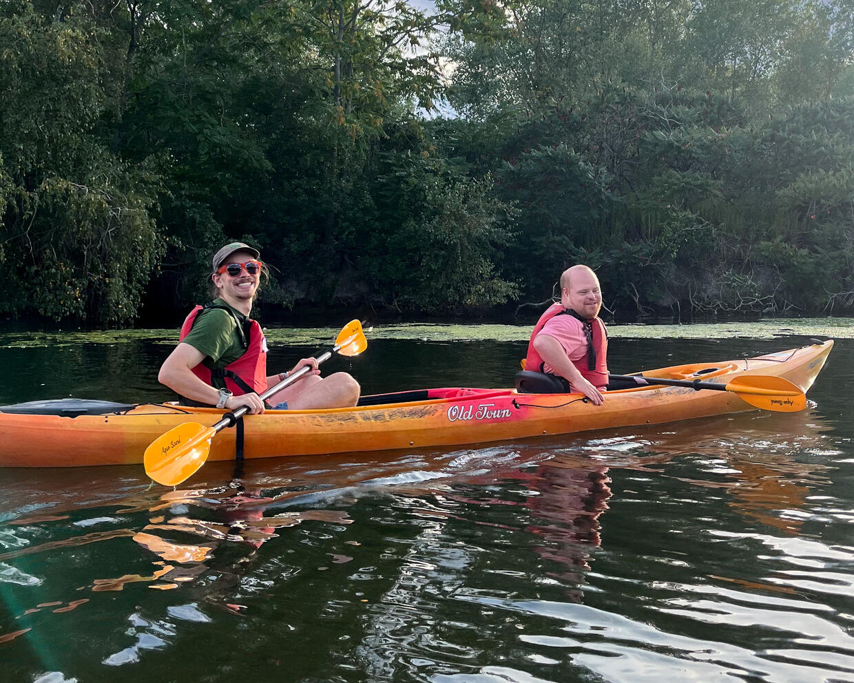 Two people paddling in a tandem kayak on a pond, smile for a photo. 