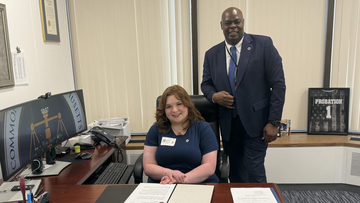 A woman sits at a desk with a man standing beside her.