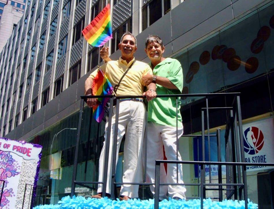Two men stand on a float. One holds a rainbow flag.