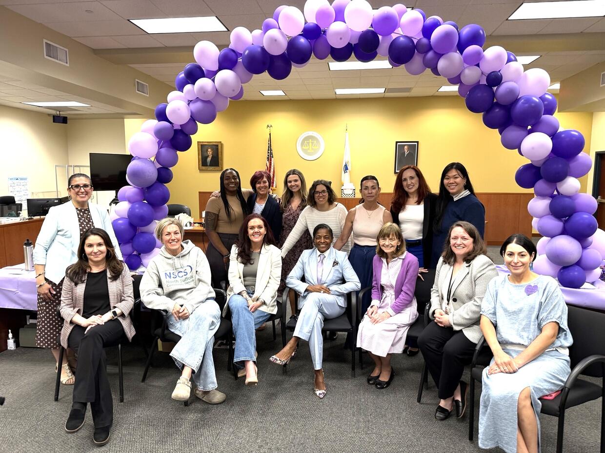 A group of women standing and seated under an arch made of purple balloons.