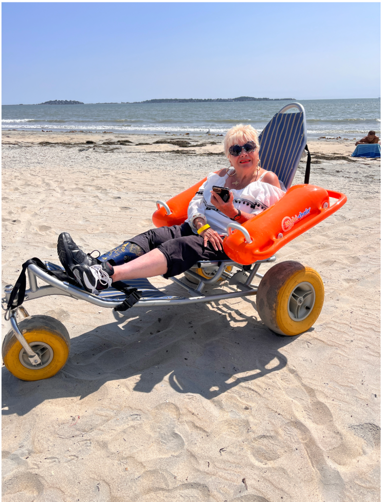 Woman with prosthetic leg smiles while seated in a beach wheelchair on the sand. 