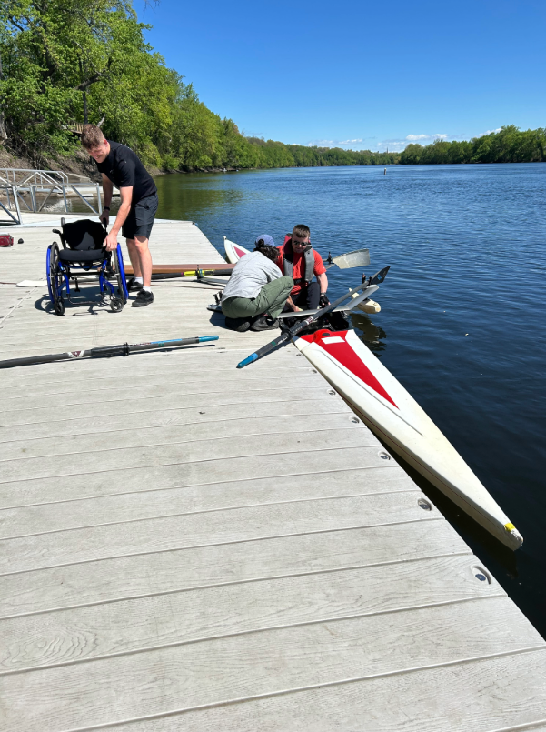 A person is assisted with adjustments while seated in a scull equipped with outriggers along a river dock. 