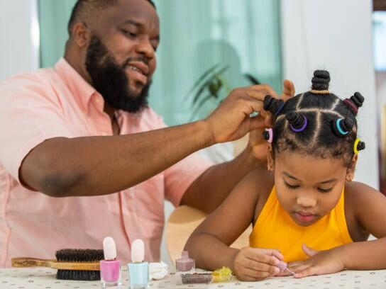 Man braiding a young girls hair, while she paints her nails with nailpolish.