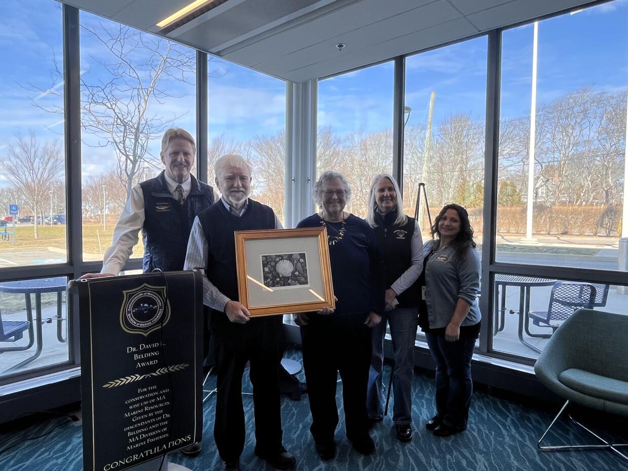 Five people stand smiling, one of the men holds a framed painting of a scallop shell. 