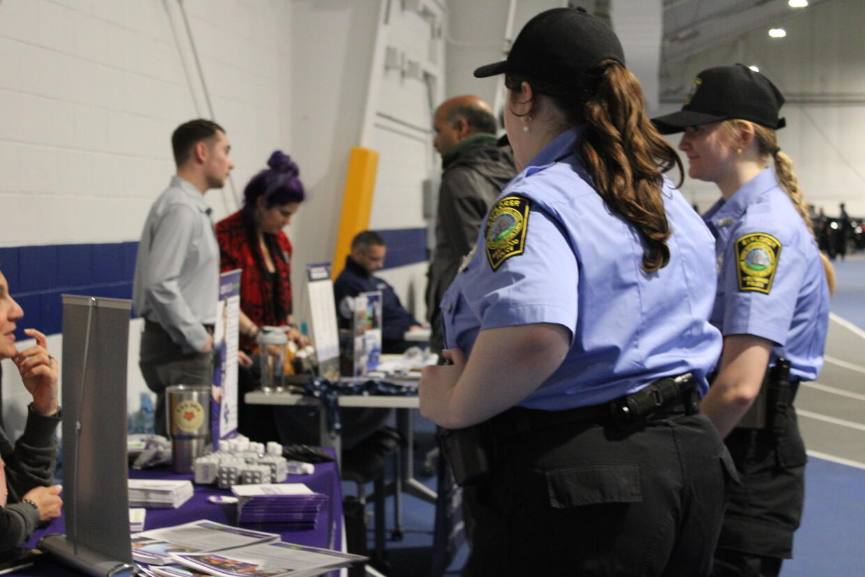 Two student cadets stand near a table with informational materials at an indoor career fair.