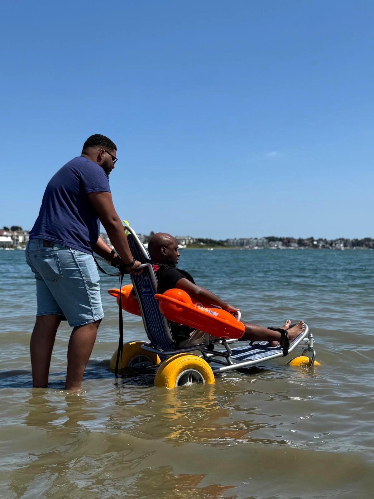A person seated in a floating beach wheelchair is pushed into the water by staff. 