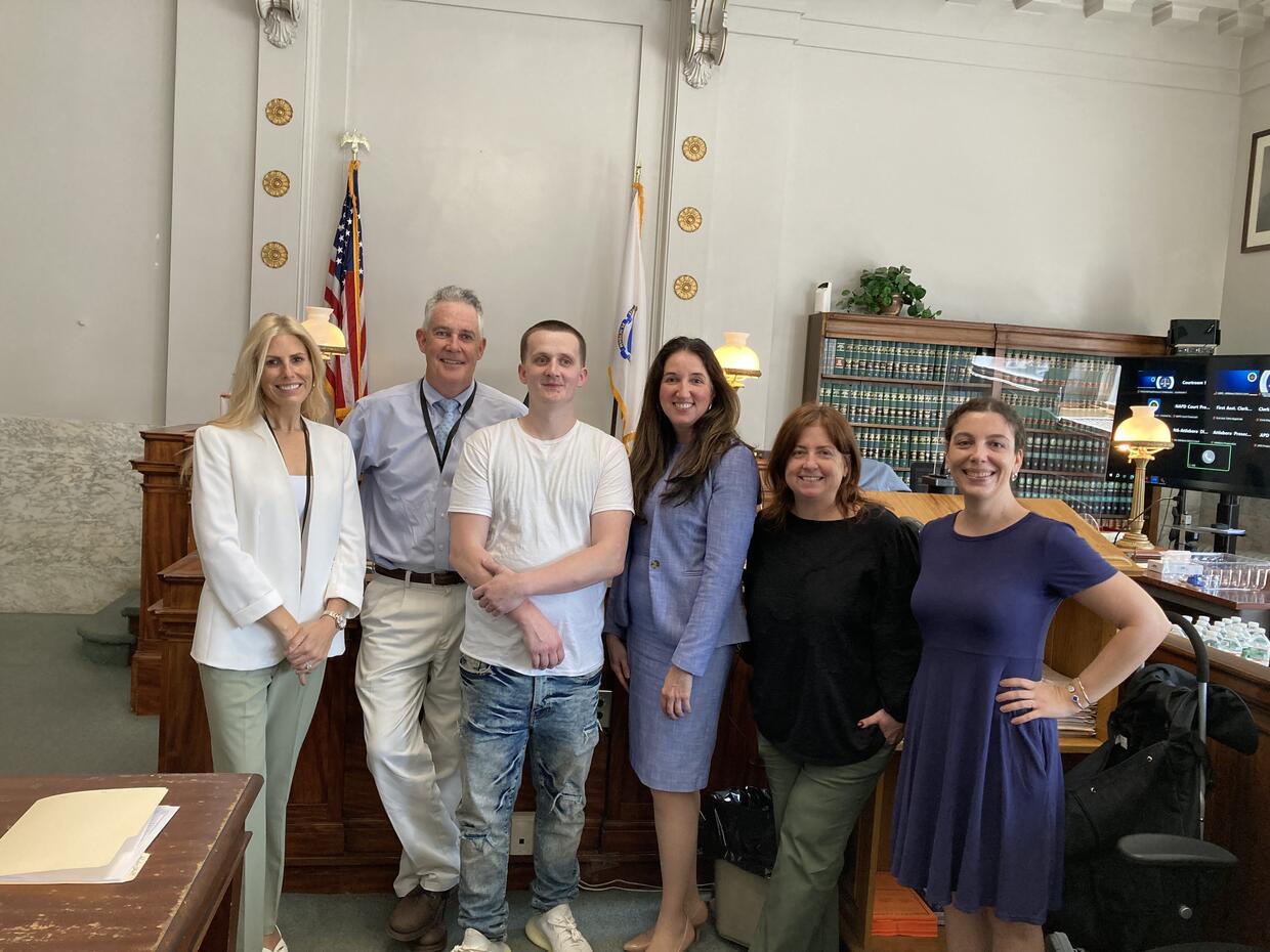 Six people standing in a courtroom with American flag, legal books, and wooden furniture visible in background.