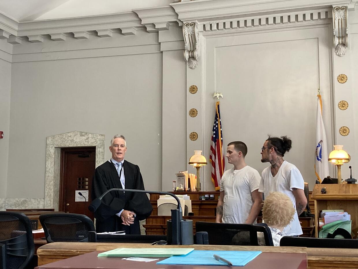 A judge stands beside two men in white shirts in a courtroom, with an American flag and courtroom decor visible in the background.