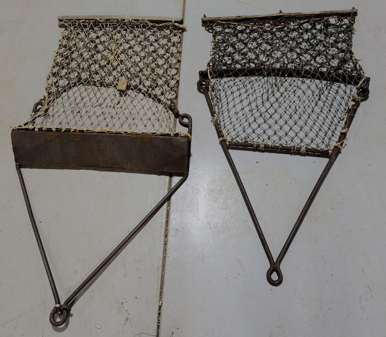 Two bay scallop dredges lay side by side, the left one has a thick bar at the opening of the net and is wider, while the right has a more narrow opening. 