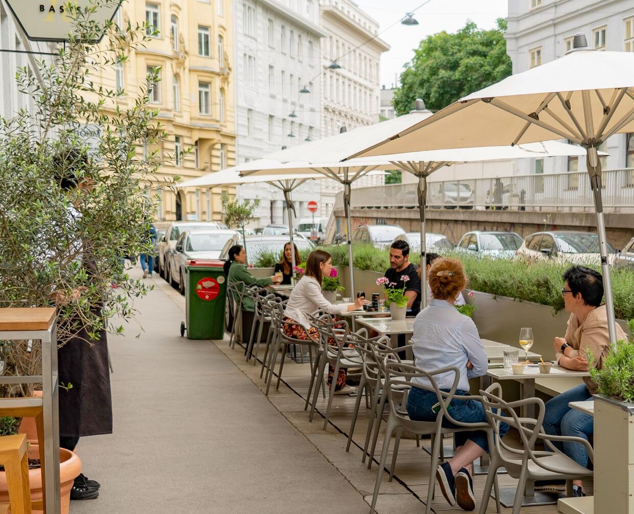 Parklet using 2-3 on-street parking spaces. There are about 6 small tables with umbrellas, with movable chairs on one side and benches on the other. The parklet is separated from the street by planters and has a wood surface.
