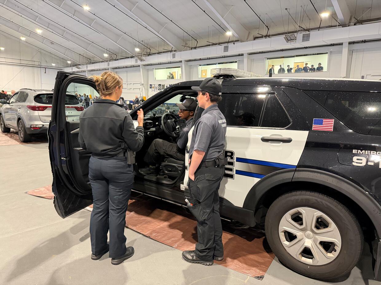 An officer stands beside a police SUV with student cadets during a station's day scenario.