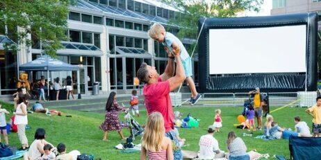 A person lifts a child while people gather for an outdoor movie screening.