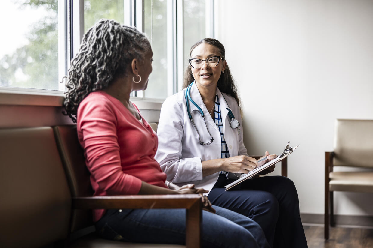 A clinician and a patient have a conversation sitting in chairs by a window. The clinician holds a clipboard and pen to make notes from the conversation.