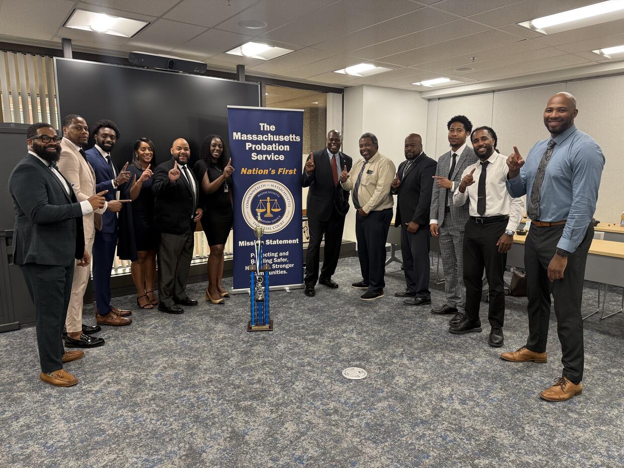 A group of eleven individuals in formal attire stands in front of a Massachusetts Probation Service banner, all raising their hands to display a "1" gesture.