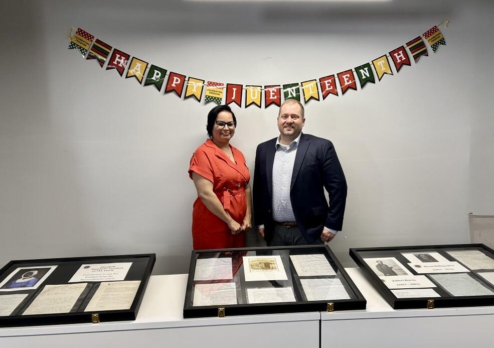 Two people standing in front of a Juneteenth banner and a table full of framed documents.