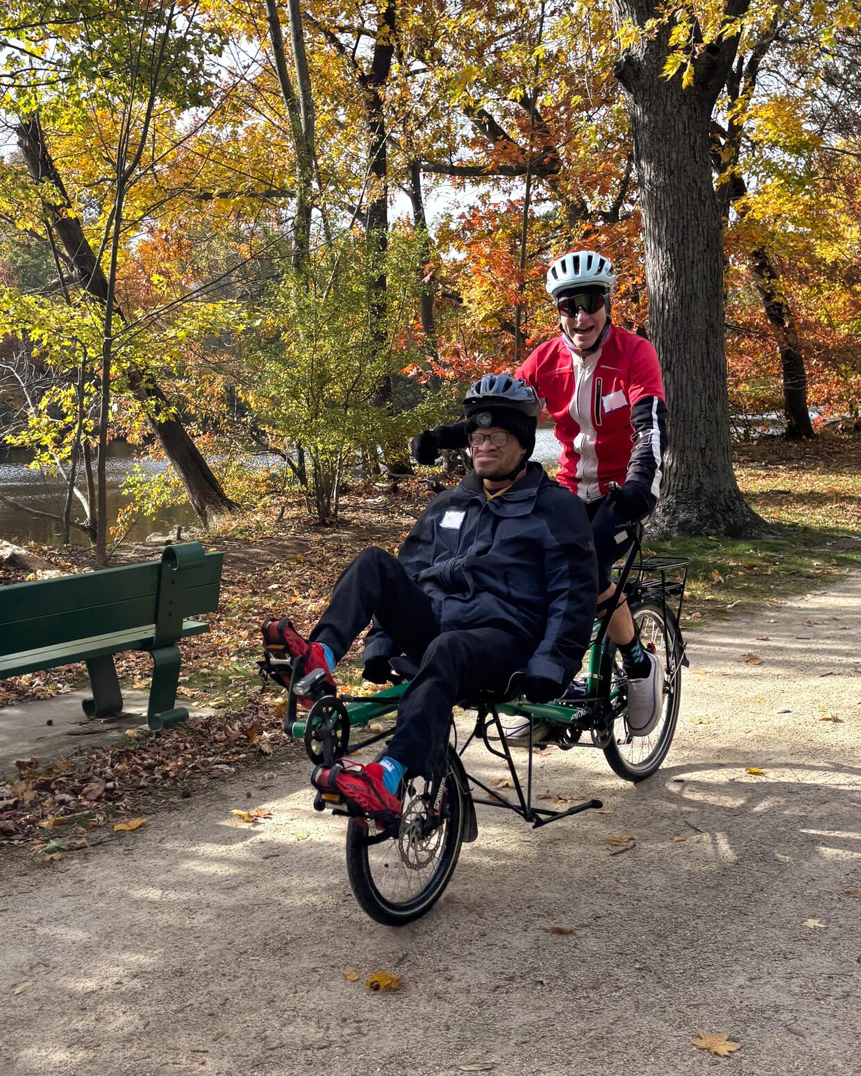 Two people are riding a semi-recumbent tandem cycle on a stone dust trail lined with autumn trees. 