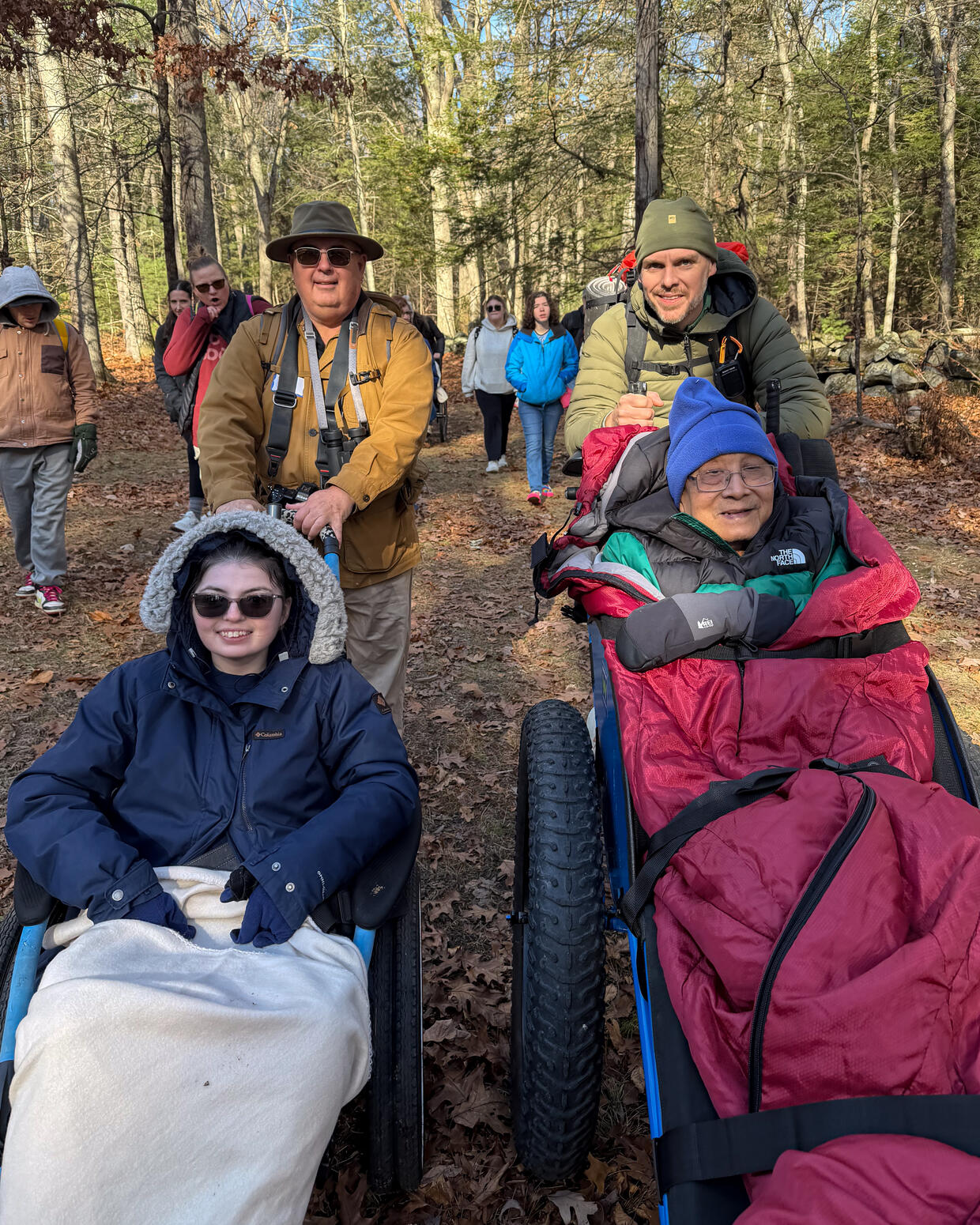 A group of people are hiking on a stone dust trail in the woods in autumn. Some are being pushed in in all-terrain wheelchairs.
