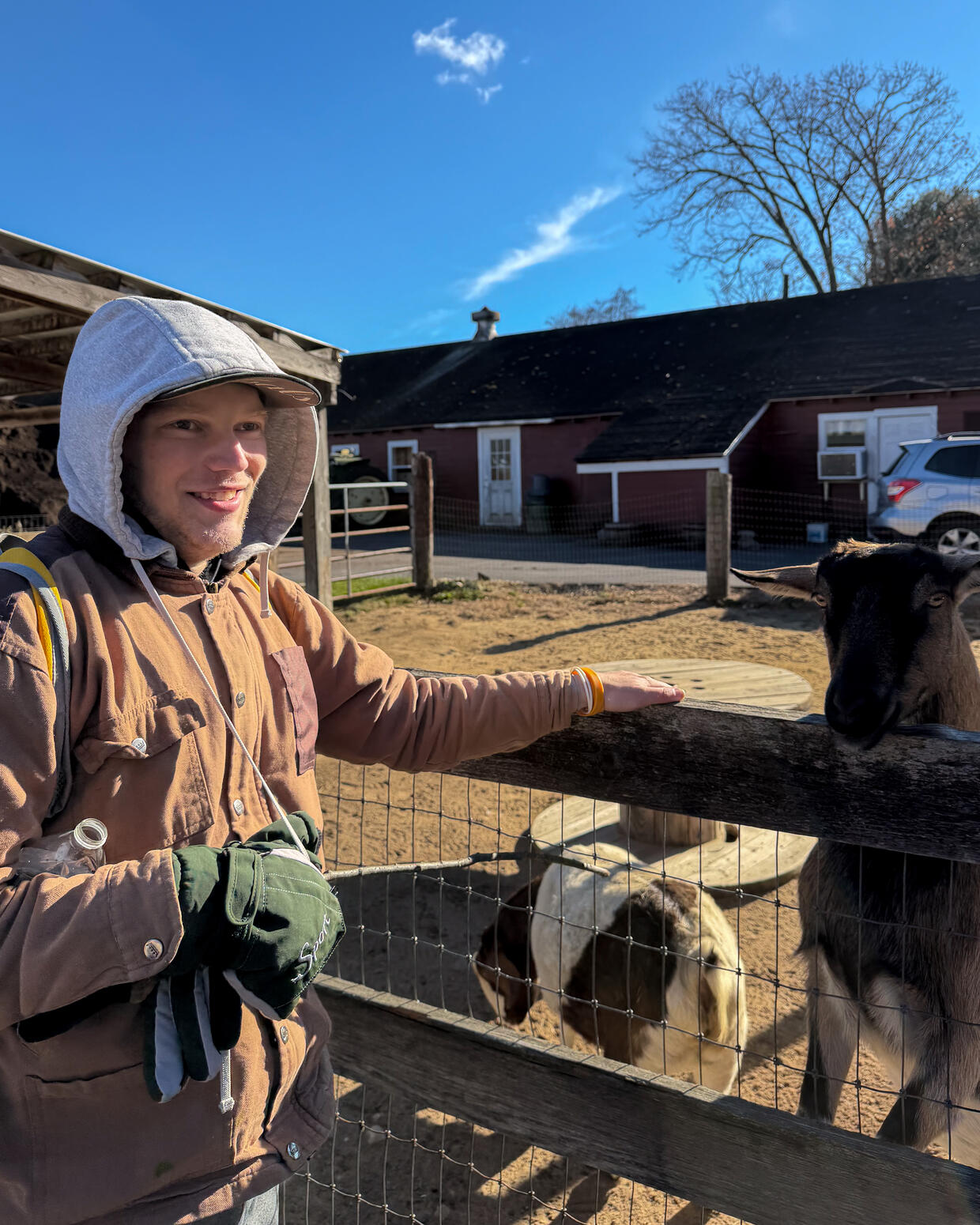 A young adult smiles while standing next to a goat pen at a farm. 