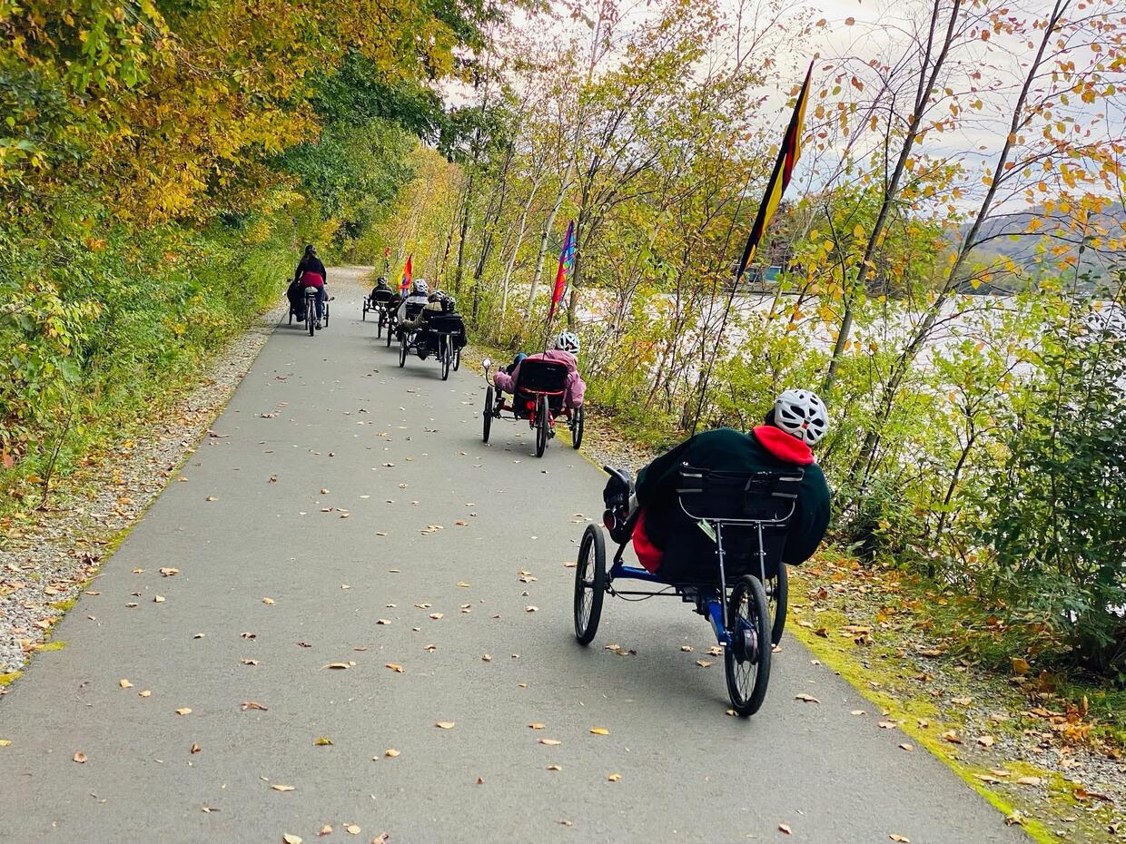 A group of people are riding recumbent trikes on a paved  trail along a lake in autumn.  