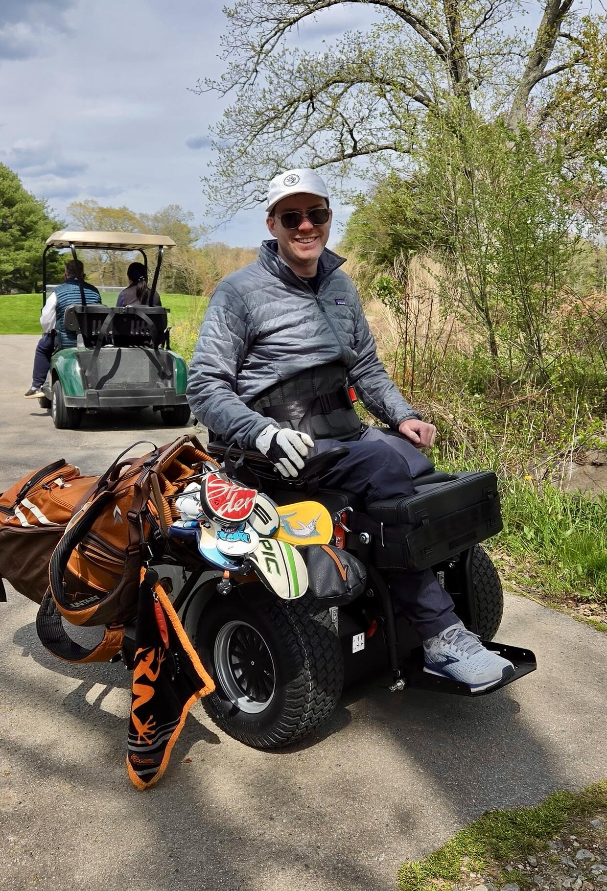 A person smiles while seated in a ParaGolfer with their golf clubs on a paved path. 