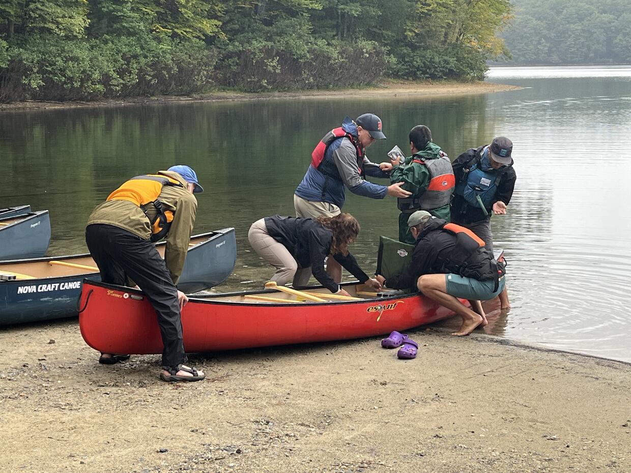 A group of people assist a participant into a canoe equipped with an adaptive seat on a lakeshore.