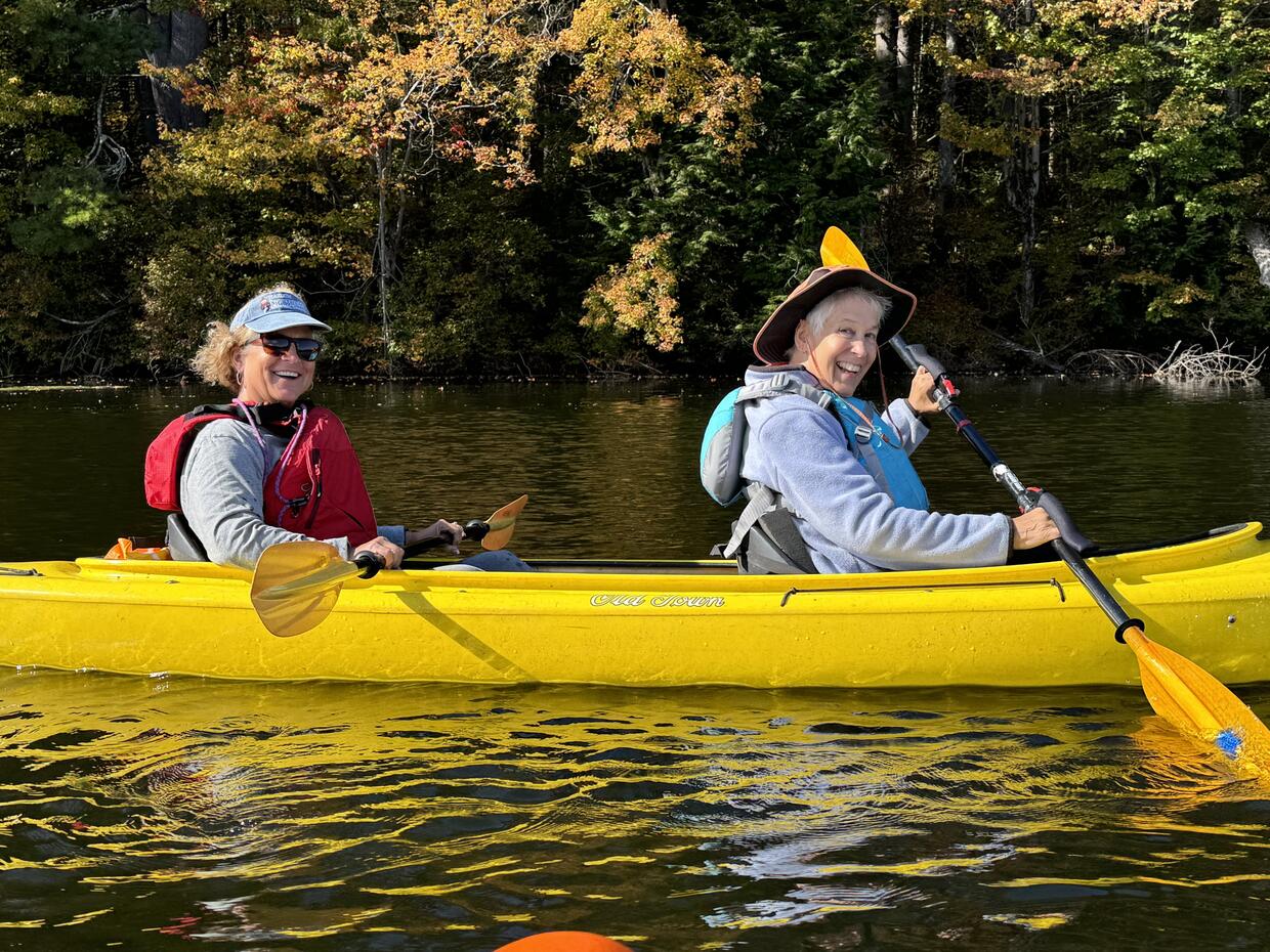 Two older adults in a tandem kayak smile while paddling on a lake in autumn. 