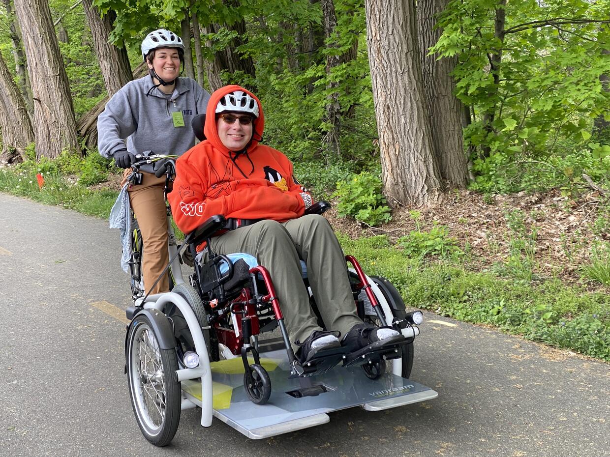 Two people are riding a wheelchair tandem cycle on a paved trail lined with trees.
