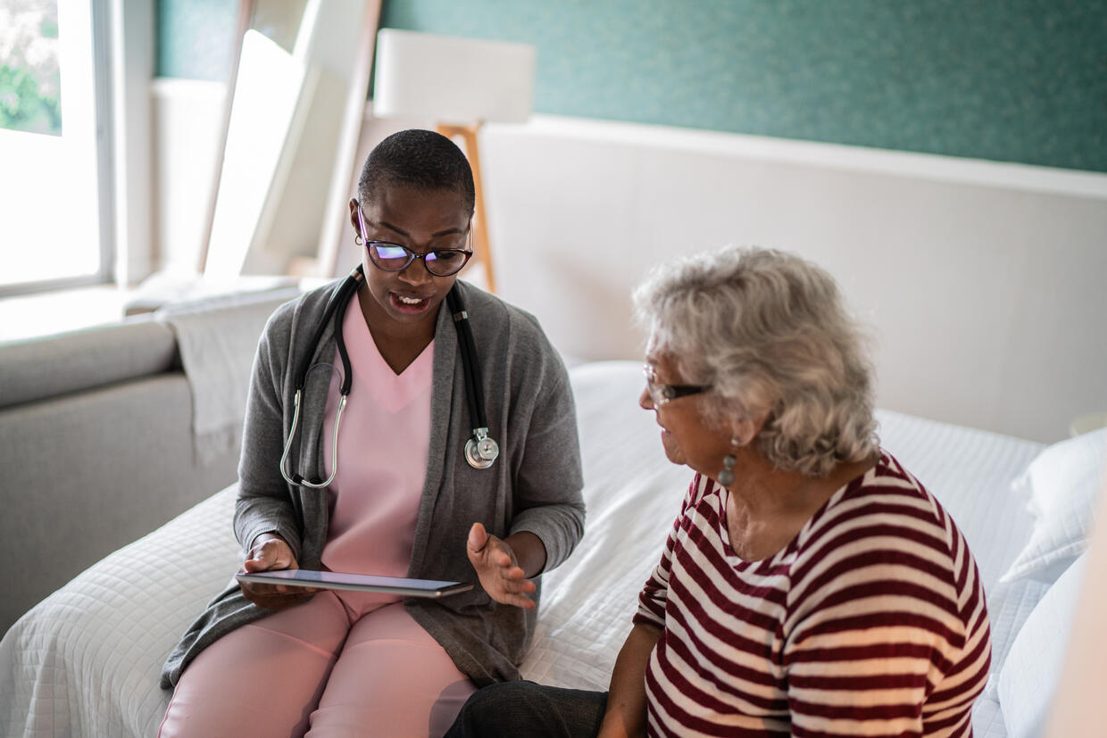 A clinician and older patient have a conversation sitting on a bed and referring to a digital handheld device. 