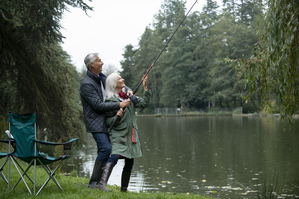 An older man and woman stand on the riverbank fishing together