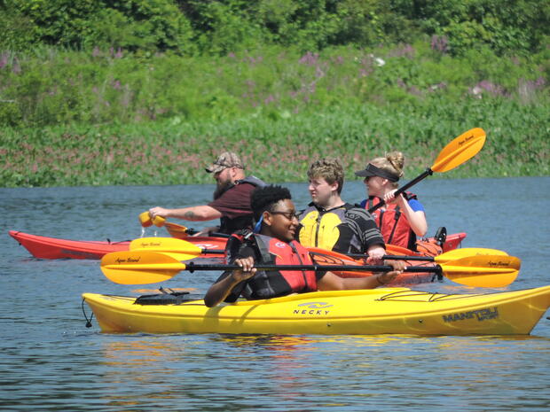 A photo of four people paddling in kayaks on the water. The shore behind the kayakers is a meadow with lush grass and flowers.