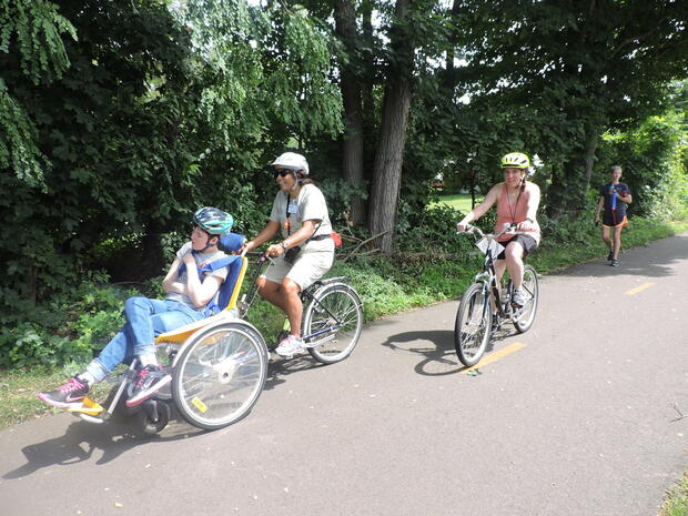 A photo of three cyclists riding together on a bike bath. The two cyclists in front are riding a tandem cycle together. The woman in the front of the tandem cycle is sitting in a seat with a back and head support. The woman behind her is pedaling and steering the cycle. The other cyclist is following on an upright bicycle.