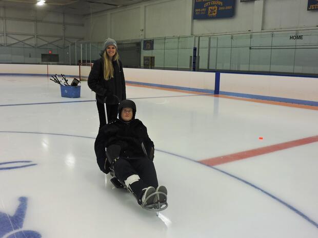 A photo of a skater sitting in an ice sled in a rink, wearing a helmet and holding a pair of sticks. Another skater is standing behind the sled and holding onto the handle. A hockey goal and hockey sticks are behind them on the ice.