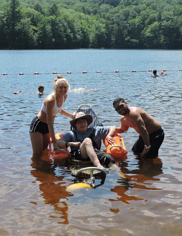 A photo of a young man sitting in a floating beach wheelchair, which is floating in the water. The chair has three wheels, and orange floats under the arm rests. Two people are standing on either side of the beach wheelchair. Everyone in the photo is smiling.