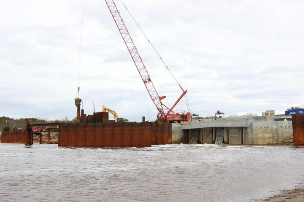 Construction equipment, including a large crane, by a bridge on a river.