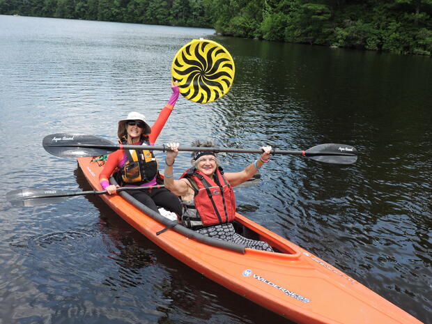 A photo of two women sitting in a tandem kayak on the water. The kayak has a large, open cockpit. The woman in front is holding up her paddle with both hands and smiling. The women behind her is holding up a large disc and also smiling.