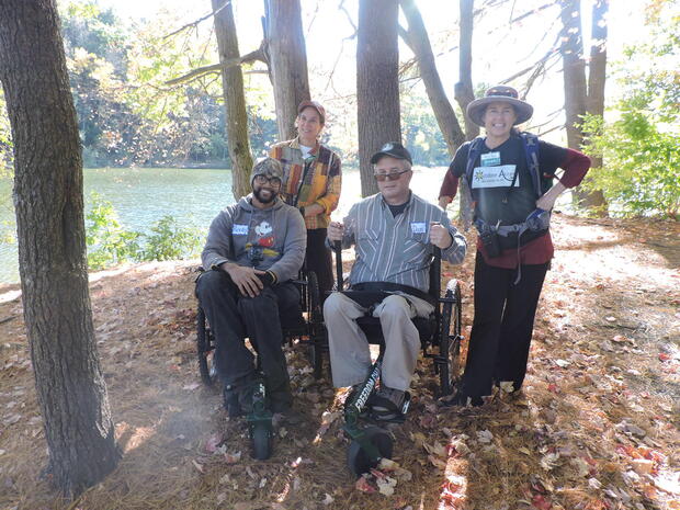 A photo of four smiling hikers in the woods next to a pond. Two of the hikers are using hiking wheelchairs, and two hikers are standing. The hiking wheelchairs have levers that the hikers push forward to move themselves.