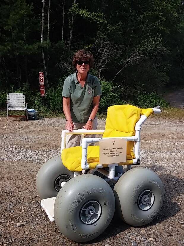 A photo of a woman in a DCR uniform standing behind a beach wheelchair and smiling. The beach wheelchair has large grey balloon tires and yellow cushions. The chair is made out of PVC pipe.
